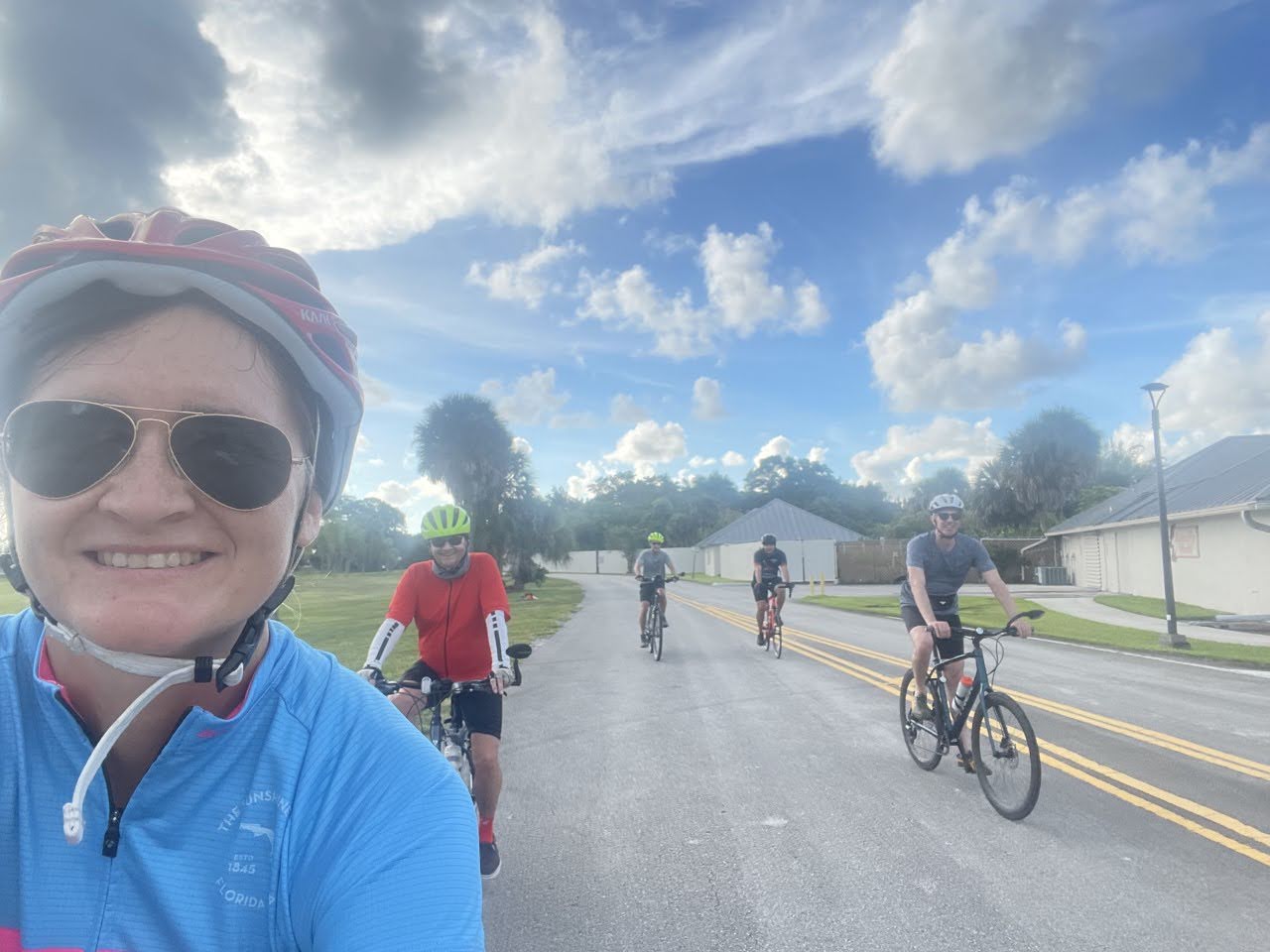 A woman takes a selfie while riding a road bike. 3 other bike riders are in the background on a sunny day. A woman takes a selfie while riding a road bike. 3 other bike riders are in the background on a sunny day.